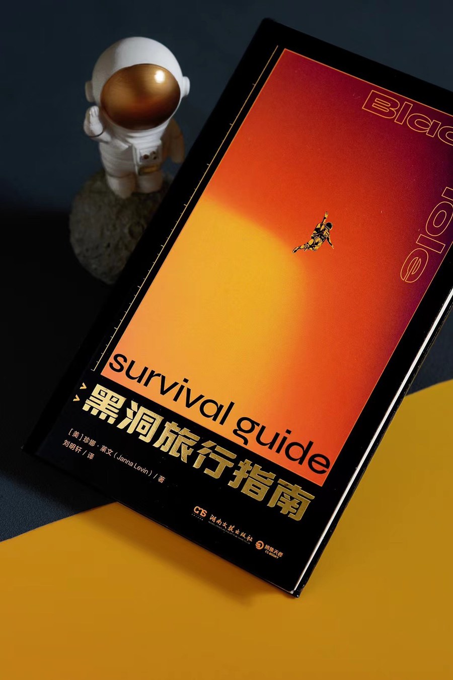 The Chinese edition of Black Hole Survival Guide standing upright, its orange cover
                showing a falling figure above the title 黑洞旅行指南, with a small astronaut figurine behind it.